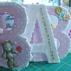 Tiny Memories-Card is a photo display shaped like the word "BABY," with eight slots for photos, designed to capture and showcase memories from a newborn's first week.