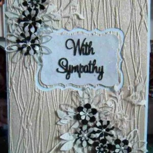 Sympathy Card-1 A cream-textured sympathy card with black and white flower embellishments and an ornate white rectangle in the centre, featuring the words "With Sympathy" in black script.