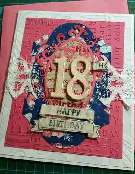 18th Birthday Card is an embossed pink background, textured white layer, and blue die-cut shape. A white lace band crosses the centre, with a peach-coloured oval frame containing a 3D number "18" and a "Happy Birthday" tag.
