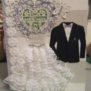 WEDDING-2 photo album is designed in the bride's chosen colours of amethyst (a deep purple) and peridot (a soft green). The front cover features the groom's black jacket and the bride's dress, which is textured with a tiered lace skirt. The dress has a shiny top, and the chest and waist are adorned with rhinestones that catch the light. At the centre of the cover, there is a heart shape made from tattered purple lace, with a smaller heart inside, made from white tattered lace. Across the heart, the words 'Mr & Mrs' are displayed, with a layer of peridot-coloured paper behind the lace, giving it a soft green glow.