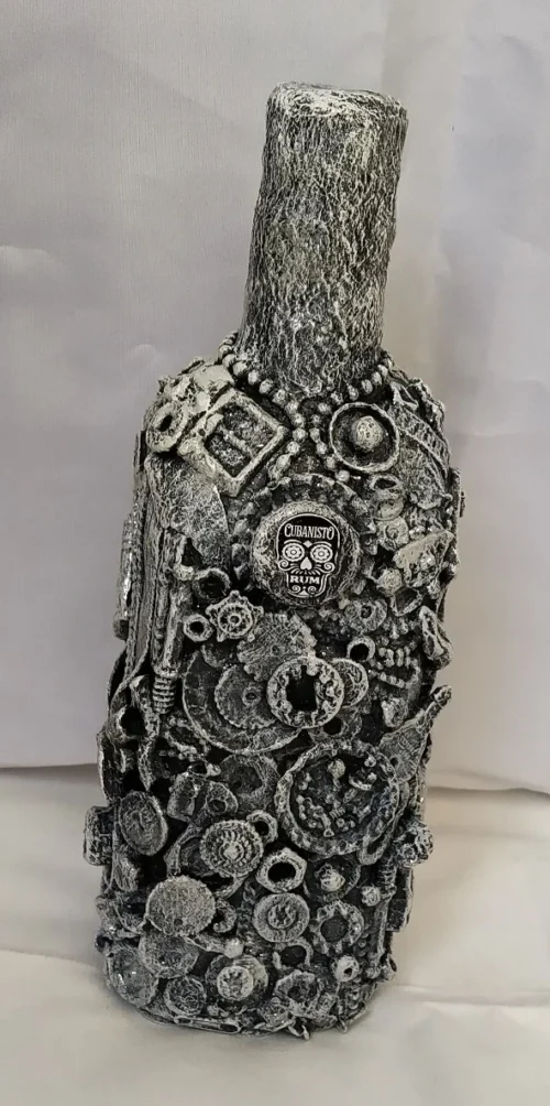 Bottle-0-Rum, front of the mixed media bottle covered by pieces of metal, plastic and textures, the background base colour is black with highlights of white on top giving the whole bottle a greyish finish.