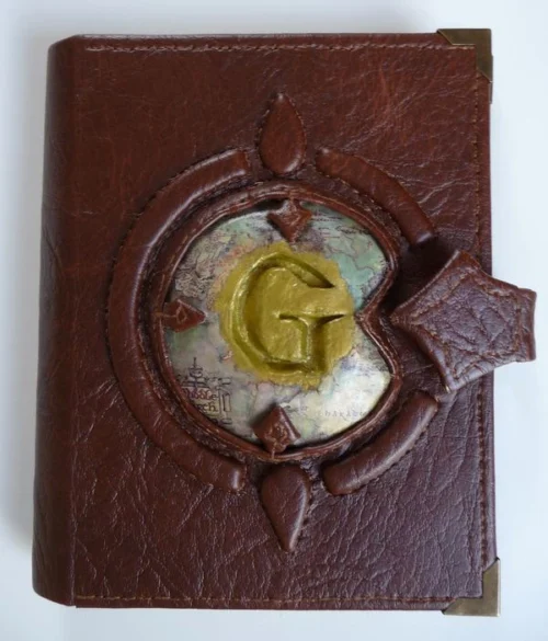 The LOR-1 sketchbook is a compact, brown faux leather-bound notebook inspired by The Lord of the Rings. It features a raised "G" on a Middle-earth map, framed by a broken circle with north and south points. The book has stitched edges, a magnetic closure, and high-quality pages for both drawing and writing. Portable and durable, it's perfect for capturing ideas on the go.