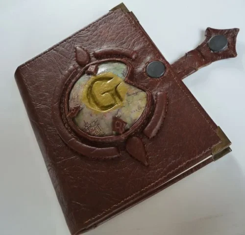 The LOR-1 sketchbook is a compact, brown faux leather-bound notebook inspired by The Lord of the Rings. It features a raised "G" on a Middle-earth map, framed by a broken circle with north and south points. The book has stitched edges, a magnetic closure, and high-quality pages for both drawing and writing. Portable and durable, it's perfect for capturing ideas on the go.